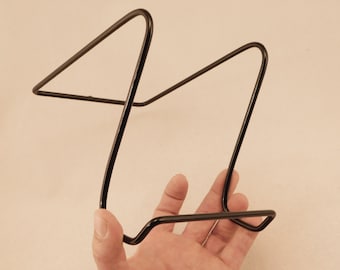 May include: Black metal book stand with a simple design. The stand is made of a single piece of wire bent into a triangular shape.