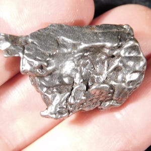May include: A small, irregularly shaped, metallic grey meteorite with a textured surface. The meteorite has a rough, uneven appearance with several indentations and protrusions. The object is held in a person's hand.