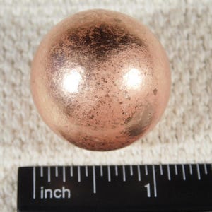 May include: A copper-colored sphere with a textured surface, approximately 1 inch in diameter, is displayed next to a ruler. The sphere has a metallic sheen and appears to be made of metal. The background is a light-colored, textured fabric.