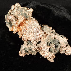 May include: A raw copper specimen with a rough, textured surface. The copper has a mix of colors, including a bright, metallic copper and areas of darker, greenish hues. The specimen is displayed against a black background.