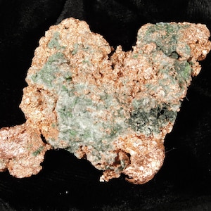 May include: A raw copper specimen with a rough, textured surface. The copper has a reddish-brown color, with patches of green and gray minerals throughout. The overall shape is irregular and organic, suggesting a natural formation.