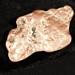 May include: A copper-colored, irregularly shaped object with a textured surface and green inclusions. The object is displayed against a dark background, highlighting its metallic sheen. The item appears to be a natural mineral specimen.