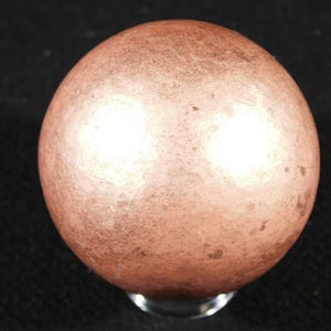 Könnte beinhalten: Ein kugelförmiges Objekt mit einer metallischen, kupferfarbenen Oberfläche, die eine texturierte, leicht verwitterte Optik aufweist. Die Kugel zeigt subtile Farbvariationen und Lichtreflexionen auf ihrer runden Form, was eine deutliche visuelle Tiefe erzeugt.