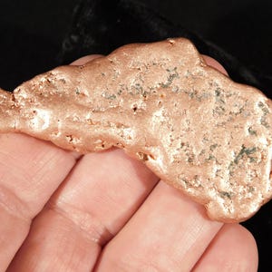 Peut inclure: Une pépite de cuivre métallique brute de forme irrégulière et allongée. La surface présente un aspect texturé et piqué avec une teinte brun-rouge chaude et de petites taches d'oxydation vert foncé dispersées sur son extérieur bosselé.