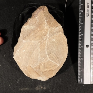 May include: A tan, teardrop-shaped stone hand axe with chipped, textured edges. A Westcott ruler is positioned vertically next to the object, showing a length of approximately 5.5 inches from the pointed tip to the rounded base.