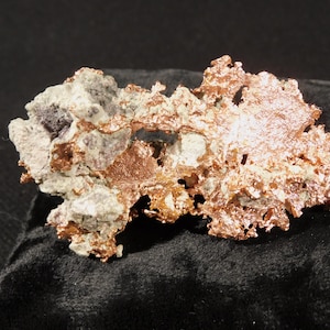 Peut inclure: Un spécimen minéral de cuivre natif brut présentant une structure cristalline métallique orange rougeâtre intégrée à des zones de matrice rocheuse gris clair et blanche, avec une texture de surface dentelée, irrégulière et poreuse.