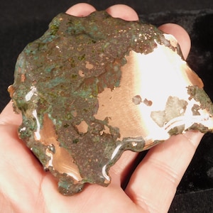 May include: A raw, irregular piece of copper metal featuring a polished, shiny metallic surface on one side and a textured, oxidized patina with shades of dark brown, green, and bronze across the remaining surface area of the object.