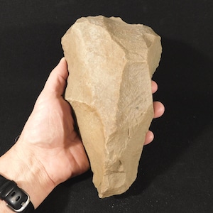 May include: A large, tan-colored stone tool with a tapered, pointed end and a wider, rounded top. The surface features multiple flat, angular facets created by flaking, resulting in a textured, uneven appearance across the entire object.