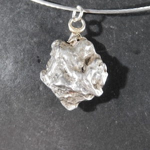May include: A silver-toned pendant with an irregular, textured, and organic shape. The pendant features a metallic, reflective surface with deep crevices and raised ridges. It is attached to a thin, circular silver-toned wire necklace by a small ring.