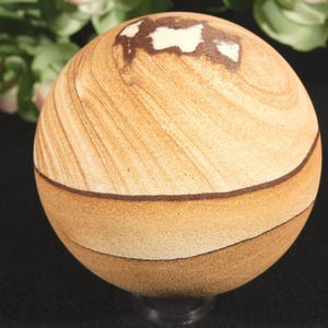 Puede incluir: Un objeto esférico de piedra con bandas horizontales naturales en tonos bronceado, beige y marrón oscuro. La superficie presenta una textura lisa y pulida con una mancha irregular de color blanco y marrón oscuro cerca del vértice superior.