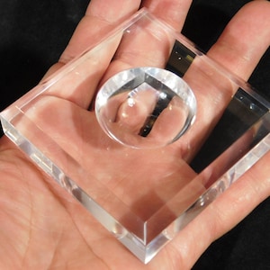Könnte beinhalten: Ein klarer, transparenter Acryl-Briefbeschwerer mit einer kreisförmigen, konvexen Linse in einem quadratischen Rahmen. Der transparente Entwurf wird in einer Hand gehalten, wodurch die polierten Kanten und die optische Klarheit hervorgehoben werden. Schlichtes, modernes Design.