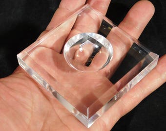 May include: A clear, transparent acrylic paperweight with a circular, convex lens set within a square frame. The paperweight is held in a hand, showcasing its polished edges and optical clarity. The design is simple and modern.