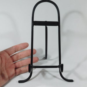 May include: Black metal easel stand for displaying artwork or photos. The stand has a simple design with a curved top and three legs.