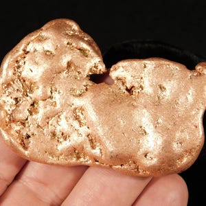 May include: A natural copper nugget with a rough, textured surface and a warm, reddish-brown color. The nugget has an irregular shape with a few small holes and a slightly shiny appearance. The background is dark.