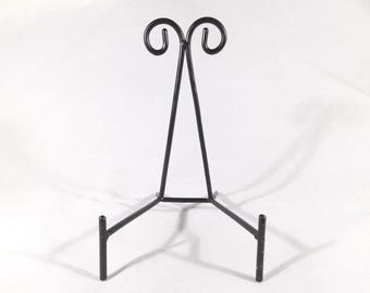 Puede incluir: Un soporte de exhibición de metal negro con una base de tres patas y un diseño decorativo de volutas en la parte superior. El soporte está diseñado para sostener un plato o una imagen. El soporte mide aproximadamente 30 cm de alto.