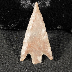 May include: A light brown arrowhead with a pointed tip and three notches at the base. The arrowhead is made of a smooth, polished stone.