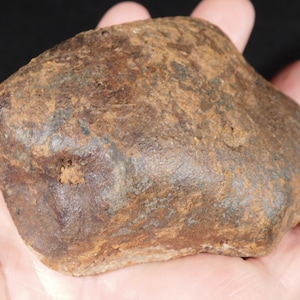 May include: A rough, irregular rock with a weathered, brownish-rust coloured surface. The stone features a dense, textured appearance with mottled dark brown and lighter tan patches across its uneven, multi-faceted exterior and a small indentation.