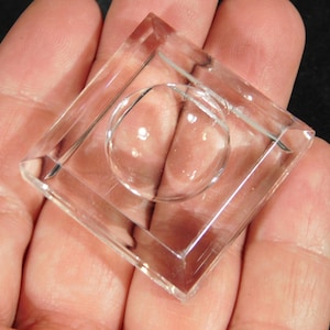 Może przedstawiać: Przezroczysty stojak wystawowy ze szkła z kwadratową podstawą i okrągłym, podniesionym środkiem. Stojak jest zaprojektowany do trzymania i prezentowania małych przedmiotów, takich jak kryształy, figurki lub inne przedmioty kolekcjonerskie.