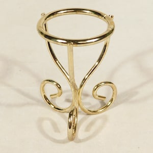 Puede incluir: Un soporte de metal dorado con una parte superior redonda y tres patas. El soporte tiene un diseño decorativo de espiral.
