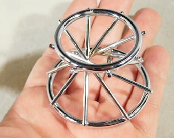 May include: A shiny, chrome-colored metal puzzle featuring two interlocking circular wheel-like structures. The puzzle is designed with spokes radiating from the center, creating a complex and intriguing design. The image showcases the puzzle's intricate details and reflective surface.
