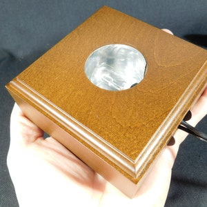 May include: A square wooden display stand with a circular cutout in the centre. The stand is a light brown colour and has a smooth finish. The stand is designed to hold a small object, such as a crystal or a figurine.