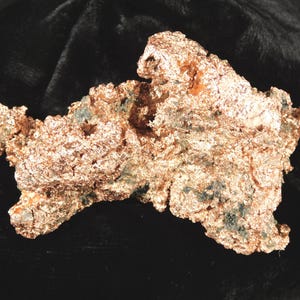 May include: A raw copper specimen with a rough, textured surface. The copper has a metallic, reddish-brown color with hints of green and blue. The shape is irregular and organic, suggesting a natural formation.