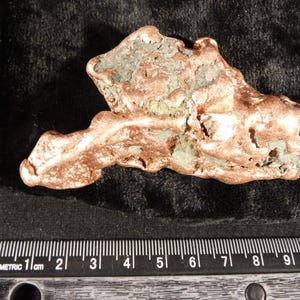 Könnte beinhalten: Ein roher, metallischer Kupferklumpen mit einer unregelmäßigen, texturierten Oberfläche in rotbraunen Farbtönen und grünen Oxidationsflecken. Ein schwarzes Lineal unter dem Objekt zeigt eine Länge von etwa 8,5 Zentimetern an.