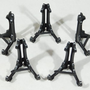 May include: Five black plastic display stands for small objects, such as figurines or photos. The stands are triangular in shape and have a simple design.