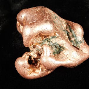May include: A raw copper nugget with a textured surface and a natural, irregular shape. The nugget displays a warm, reddish-brown color with hints of green and sits against a dark background. This item is a natural mineral specimen.