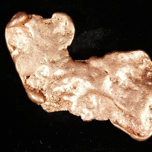 May include: A natural, irregular-shaped gold nugget with a textured surface and a warm, metallic rose gold color. The nugget is displayed against a black background, highlighting its unique form and luster. A piece of raw gold.