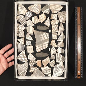 May include: A collection of ancient pottery shards with geometric patterns in a rectangular box. The shards are primarily gray and white, with various designs including crosshatching and grid patterns. A ruler is placed next to the box for scale.