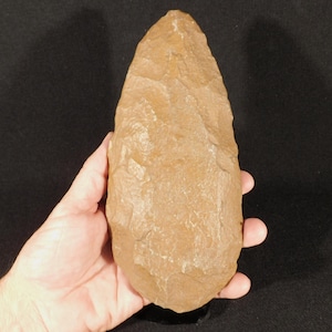 May include: A tan-colored, teardrop-shaped stone tool featuring a pointed tip and a rounded base. The surface displays distinct, overlapping flake scars across its entire body, indicating a knapped construction with a textured, matte finish.