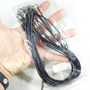 May include: A clear plastic bag filled with multiple black waxed cord necklaces. Each necklace has a silver-coloured clasp. The cords are approximately 43 cm long. The bag is held in a person's hand.