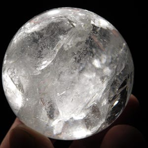 May include: A clear quartz crystal sphere, held against a black background. The sphere is translucent with internal fractures and inclusions, creating a textured appearance. The light catches the facets, giving it a sparkling effect.