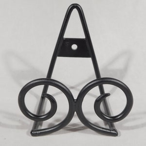 May include: Black metal display stand with a decorative scroll design. The stand is shaped like an A and has a small hole in the top.