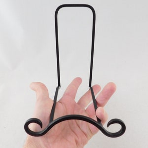 May include: Black metal plate display stand with a curved base and a vertical support. The base features decorative scrollwork. The stand is designed to hold and display plates or other decorative items.
