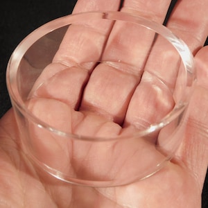 May include: A clear acrylic ring holder, shown in a person's hand. The ring holder is circular and has a smooth, polished finish.