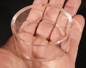 May include: A clear acrylic ring holder, shown in a person's hand. The ring holder is circular and has a smooth, polished finish.