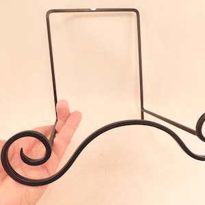May include: Black metal display stand with a decorative scroll design. The stand is designed to hold plates, photos, or other items.