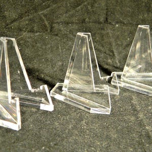 May include: Three clear acrylic display stands. Each stand has a triangular support and a rectangular base. The stands are designed to hold small items, such as collectibles or artwork. The stands are transparent and allow for a clear view of the displayed items.