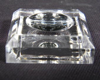 May include: A clear glass display stand with a square base and a raised circular platform. The stand is designed to showcase small objects.