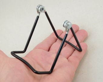 May include: A black metal stand with two adjustable legs. The stand is designed to hold a small object, such as a phone or a book.