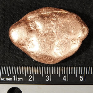 May include: A copper-colored, irregularly shaped nugget, possibly a mineral specimen, sits on a dark surface. A metric ruler is placed below the nugget, showing measurements in centimeters. The nugget appears to be approximately 5 cm in length.