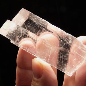 May include: A clear, rectangular selenite crystal specimen with visible internal fractures and a smooth, translucent surface. The mineral displays a distinct geometric shape with sharp edges and a glassy, reflective quality that catches light.