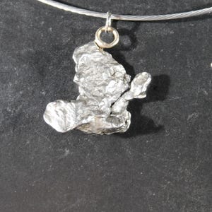 May include: A silver-toned, irregularly shaped metallic pendant with a rough, textured surface. The pendant is suspended from a thin, circular silver-coloured wire necklace by a small, round metal jump ring, creating a simple and distinct piece.
