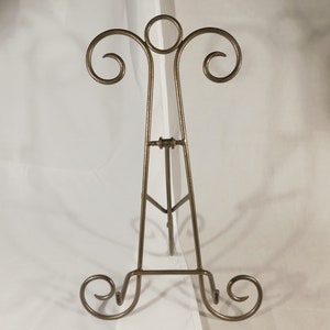 May include: A bronze-colored metal easel with a decorative design. The easel has a three-legged base and a top with a circular element and scrolled accents. The easel is designed to hold a picture frame or artwork.