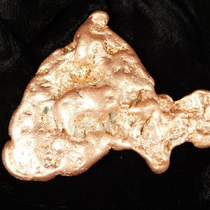 May include: A raw copper nugget with a textured surface and a natural, irregular shape. The nugget has a warm, reddish-brown color and appears to be unpolished. The background is a dark, solid color.
