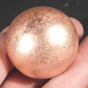 May include: A copper-colored sphere with a textured surface. The sphere is held in a hand, and the metallic surface reflects light. The overall color is a warm, reddish-brown.