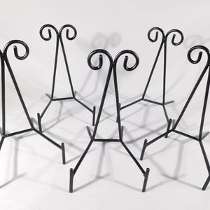 May include: Six black metal easel stands with a decorative scroll design at the top. The stands are arranged in a circle.