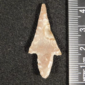 Puede incluir: Una punta de flecha antigua de color marrón claro, con una base dentada y una punta afilada. La punta de flecha de piedra se encuentra sobre un fondo negro. Se puede ver una regla con marcas en centímetros.
